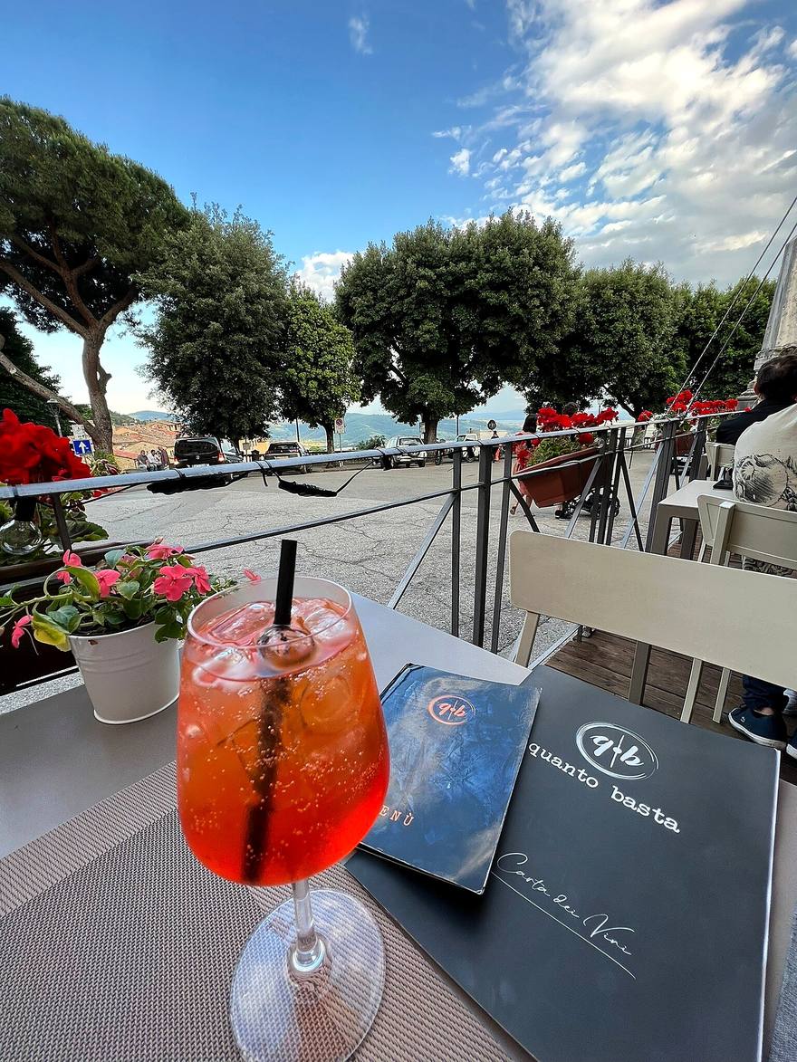 Aperol Spritz sulla terrazza accanto al menù con il marchio q·b, con gerani rosa e i pini sulla Valdichiana